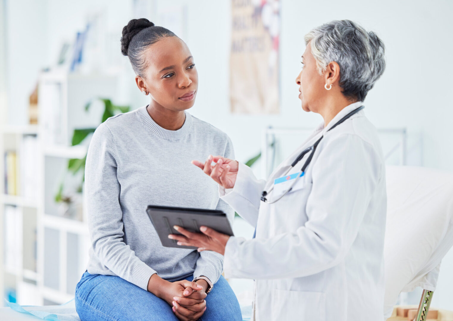 A doctor with gray hair discusses something on a tablet with a sitting woman in a gray sweater in a medical office.