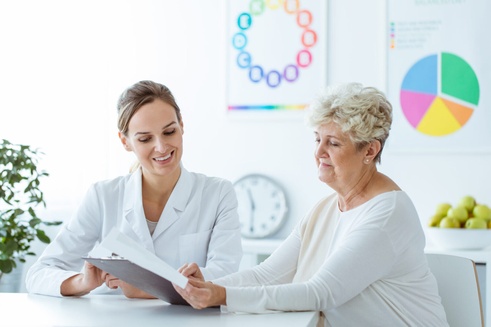 A doctor discusses a clipboard with an elderly woman in a bright, modern office with colorful charts and a bowl of apples visible in the background.